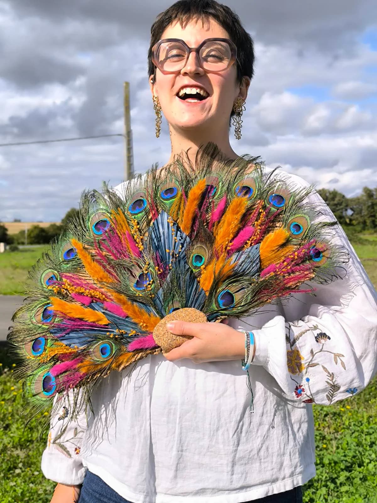 Cathy souriante, tenant une composition en éventail de plumes de paon et de fleurs séchées colorées