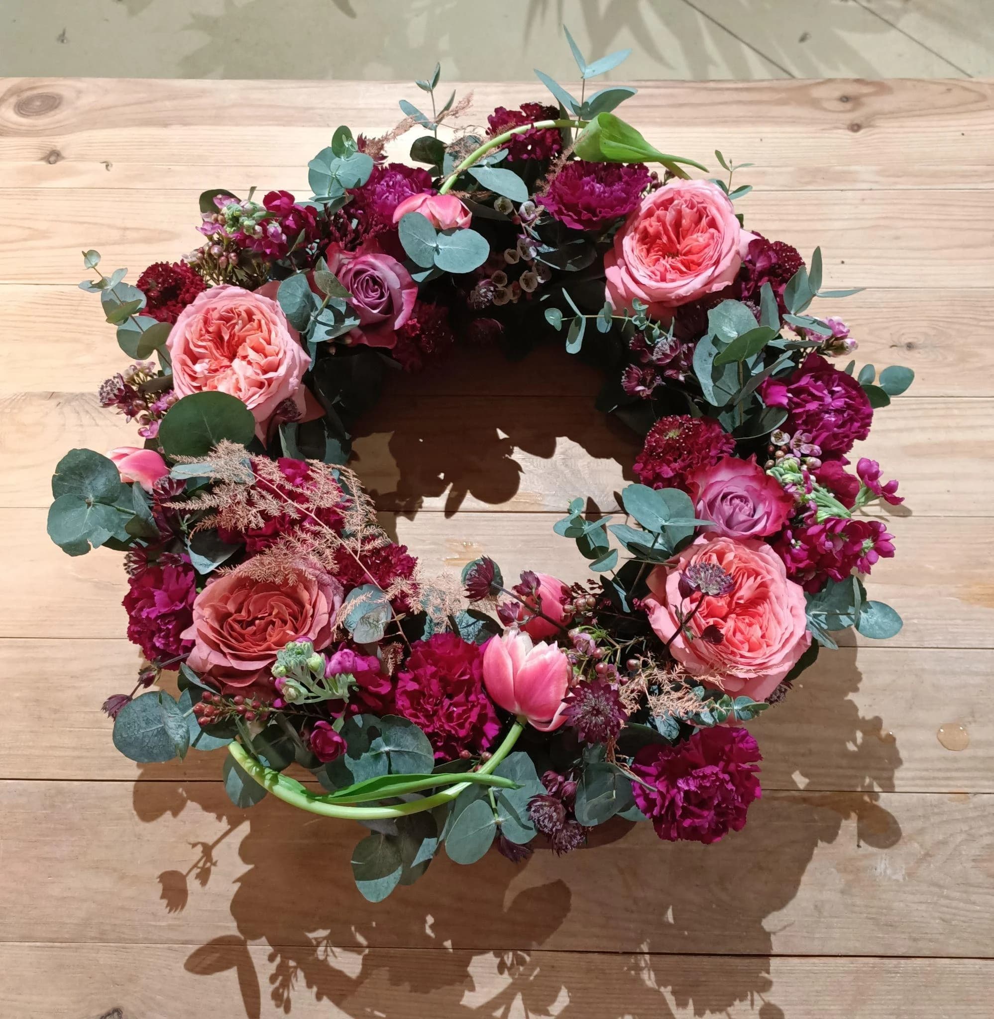 Couronne de deuil généreuse composée de roses, tulipes et fleurs fuchsia, prune et rose sur fond de bois